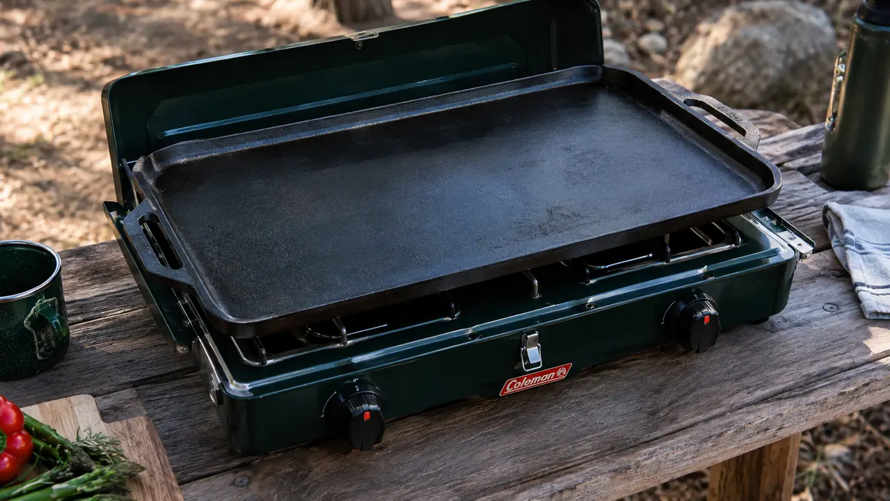 A two-burner camp stove with a large cast iron griddle centered over both burners to make a single wide cooking surface